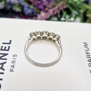 May include: A silver ring with five clear gemstones set in a row. The ring is on a white surface with the word "PARIS" visible. The ring's design features intricate detailing on the band.