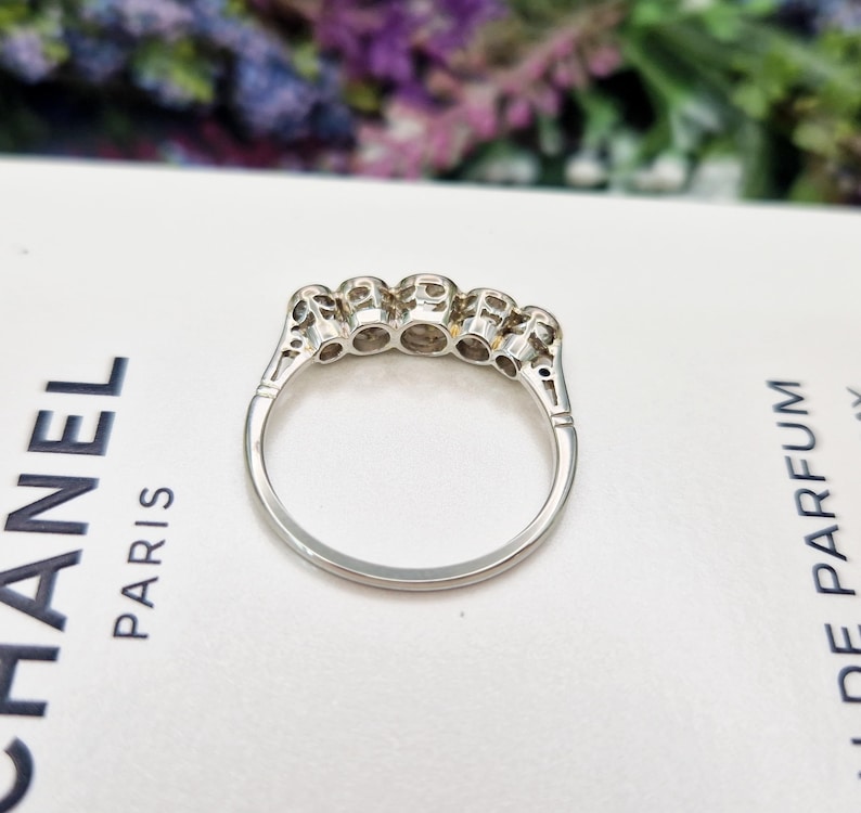 May include: A silver ring with five clear gemstones set in a row. The ring is on a white surface with the word "PARIS" visible. The ring's design features intricate detailing on the band.