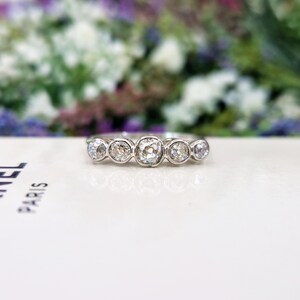 May include: A silver ring with five round, clear gemstones. The ring is set on a white surface with a blurred floral background. The gemstones are of varying sizes, with the center stone being the largest.