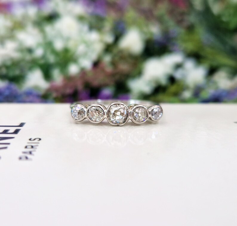 May include: A silver ring with five round, clear gemstones. The ring is set on a white surface with a blurred floral background. The gemstones are of varying sizes, with the center stone being the largest.