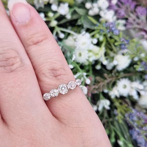 May include: A silver ring with seven round, clear gemstones of varying sizes. The ring is set on a finger, with a blurred floral background of white, purple, and green.