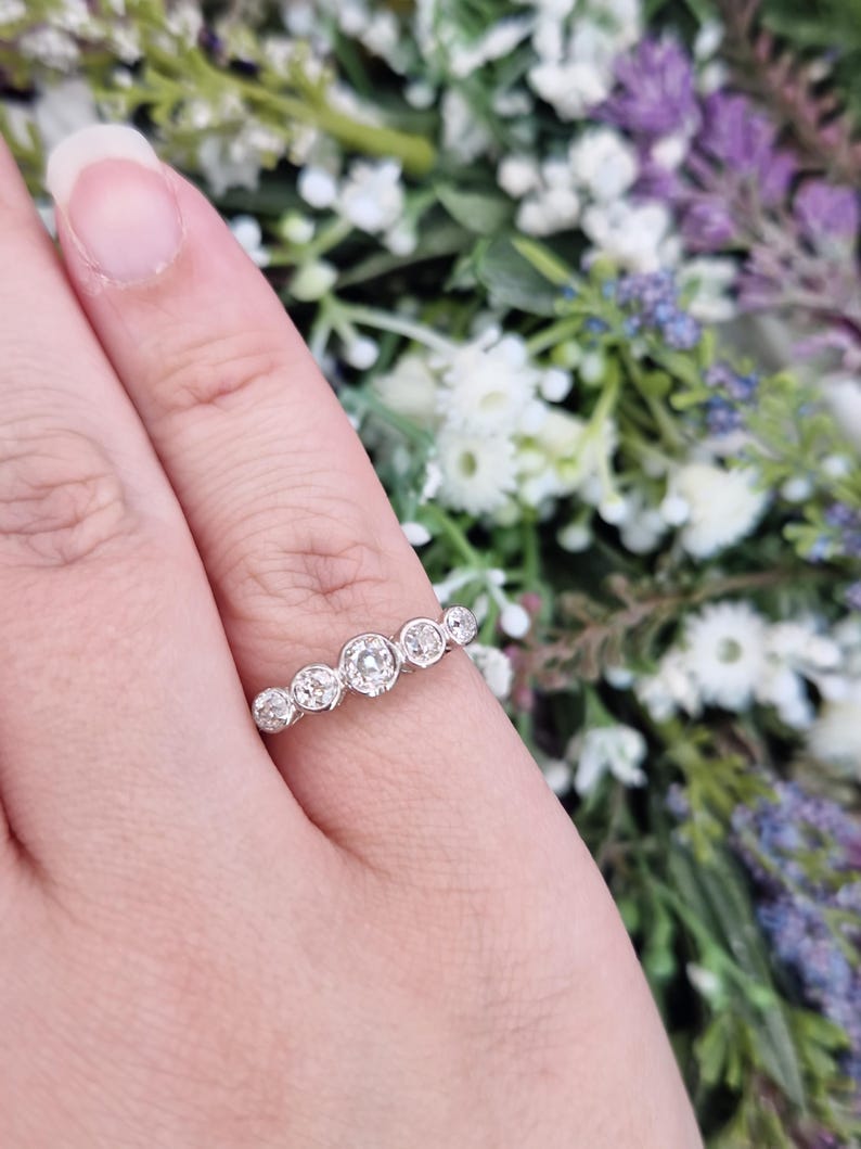 May include: A silver ring with seven round, clear gemstones of varying sizes. The ring is set on a finger, with a blurred floral background of white, purple, and green.