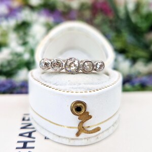 May include: A silver ring with five round, clear diamonds of varying sizes, set in a white jewelry box. The ring is centered in the box, with the diamonds facing up. The box is white with gold accents and a gold clasp.