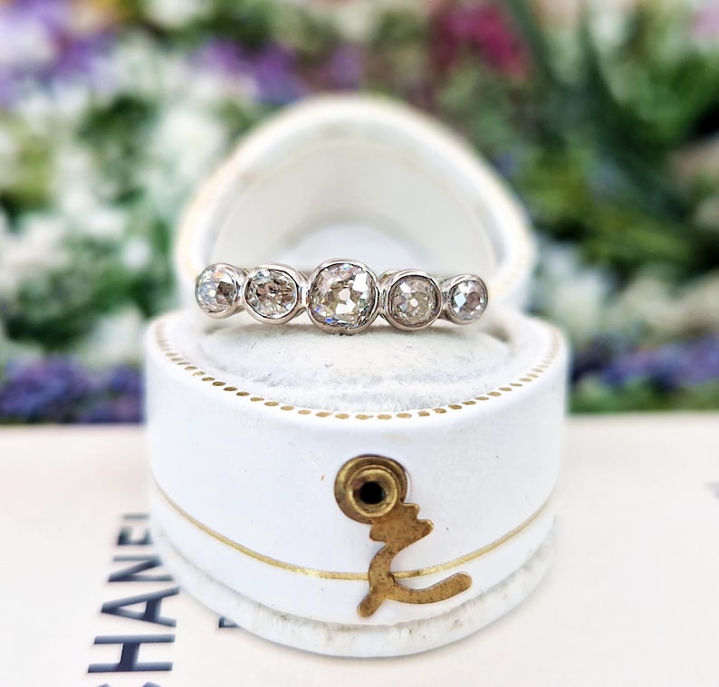 May include: A silver ring with five round, clear diamonds of varying sizes, set in a white jewelry box. The ring is centered in the box, with the diamonds facing up. The box is white with gold accents and a gold clasp.