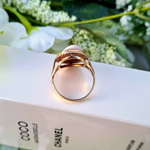 Op de afbeelding: Een gouden ring met een roze ovale edelsteen gezet in een gedetailleerde zetting. De ring ligt op een wit oppervlak.