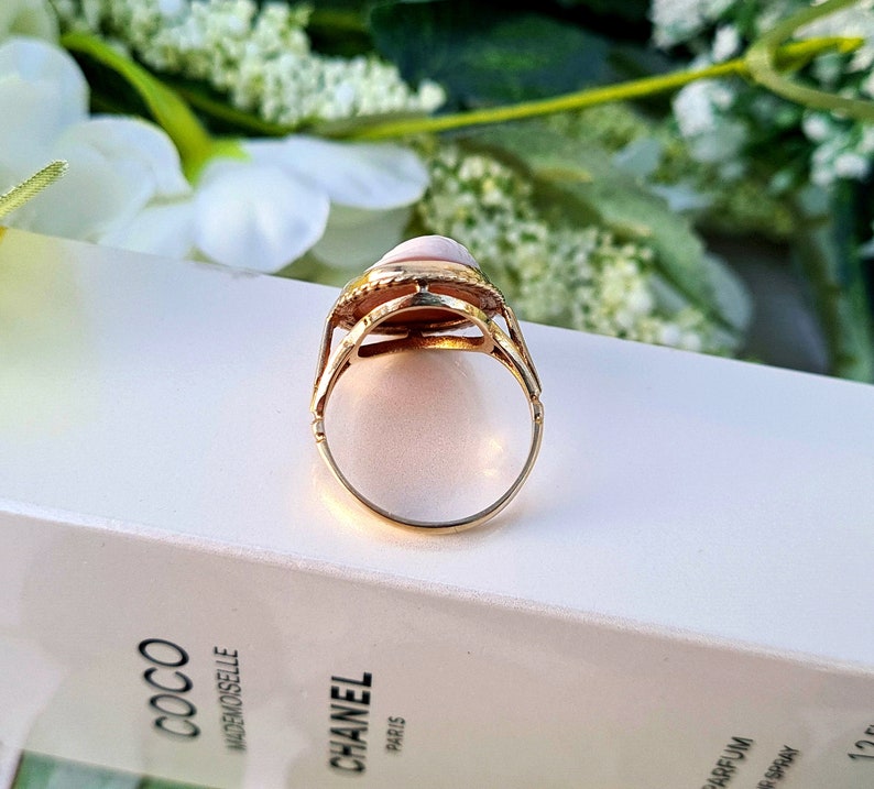 Op de afbeelding: Een gouden ring met een roze ovale edelsteen gezet in een gedetailleerde zetting. De ring ligt op een wit oppervlak.