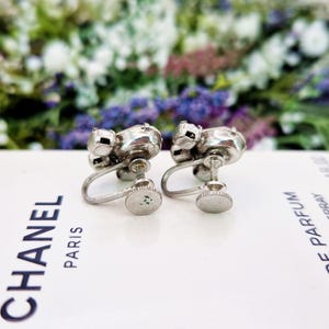 May include: A pair of silver tone clip-on earrings with clear rhinestones. The earrings are in the shape of a flower with five petals.
