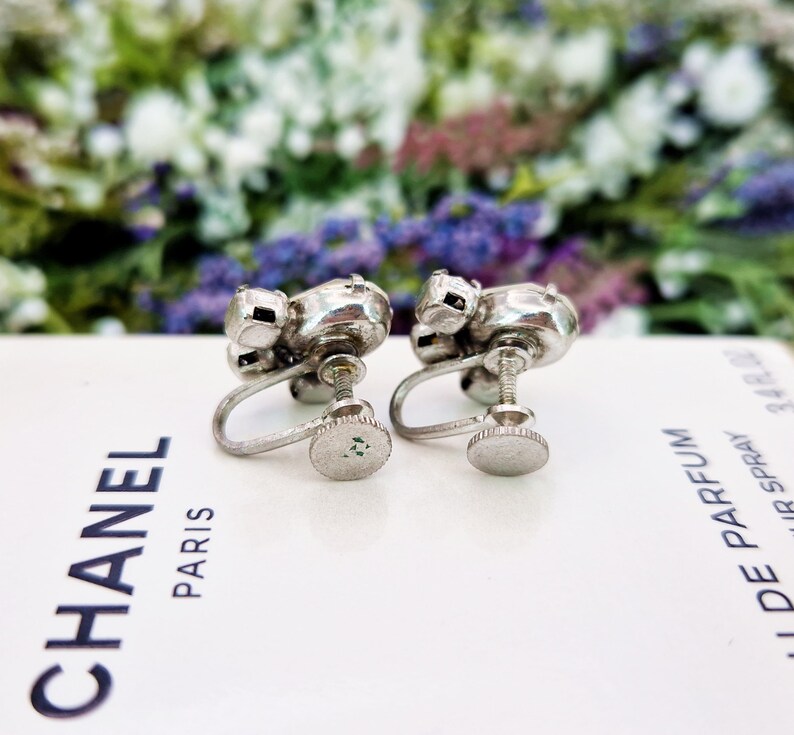 May include: A pair of silver tone clip-on earrings with clear rhinestones. The earrings are in the shape of a flower with five petals.