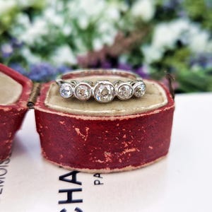 May include: A silver ring with five round, clear diamonds set in individual bezels. The ring is displayed inside a vintage red jewelry box with a cream-colored interior. The background features blurred greenery and flowers.