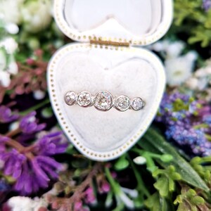 May include: A silver ring with five round, clear diamonds is displayed inside a heart-shaped, white jewelry box. The ring is set against a backdrop of colorful flowers, including purple and white blooms.
