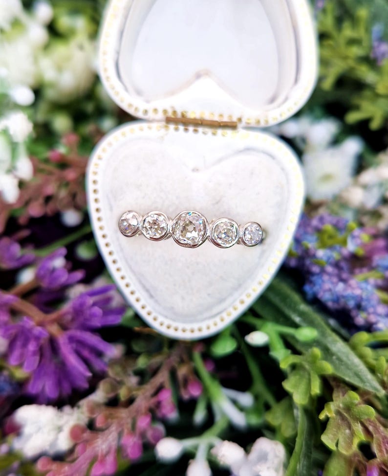 May include: A silver ring with five round, clear diamonds is displayed inside a heart-shaped, white jewelry box. The ring is set against a backdrop of colorful flowers, including purple and white blooms.