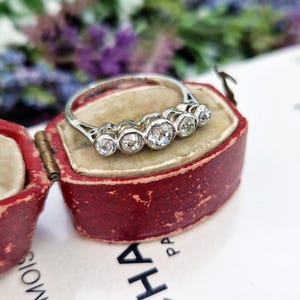 May include: A silver ring with five clear, round gemstones, displayed in a vintage red jewelry box. The ring's band is thin and delicate, and the gemstones are set close together. The box is open, revealing the ring against a soft, worn interior.