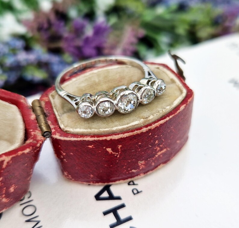 May include: A silver ring with five clear, round gemstones, displayed in a vintage red jewelry box. The ring's band is thin and delicate, and the gemstones are set close together. The box is open, revealing the ring against a soft, worn interior.