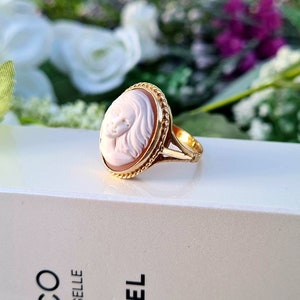 Op de afbeelding: Een gouden ring met een cameo met een gegraveerd portret van een vrouwengezicht. De cameo is gezet in een ovale bezel met een touwachtige rand. De ring ligt op een wit oppervlak.