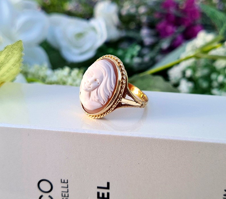 Op de afbeelding: Een gouden ring met een cameo met een gegraveerd portret van een vrouwengezicht. De cameo is gezet in een ovale bezel met een touwachtige rand. De ring ligt op een wit oppervlak.