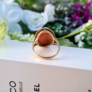 Op de afbeelding: Een gouden ring met een roze ovale edelsteen in een bezel-setting. De ring ligt op een wit oppervlak.