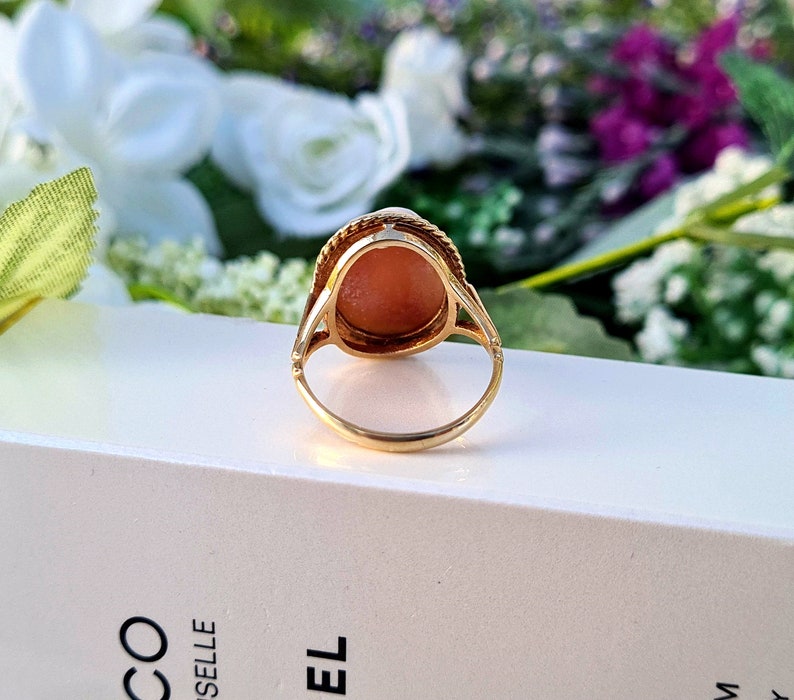 Op de afbeelding: Een gouden ring met een roze ovale edelsteen in een bezel-setting. De ring ligt op een wit oppervlak.