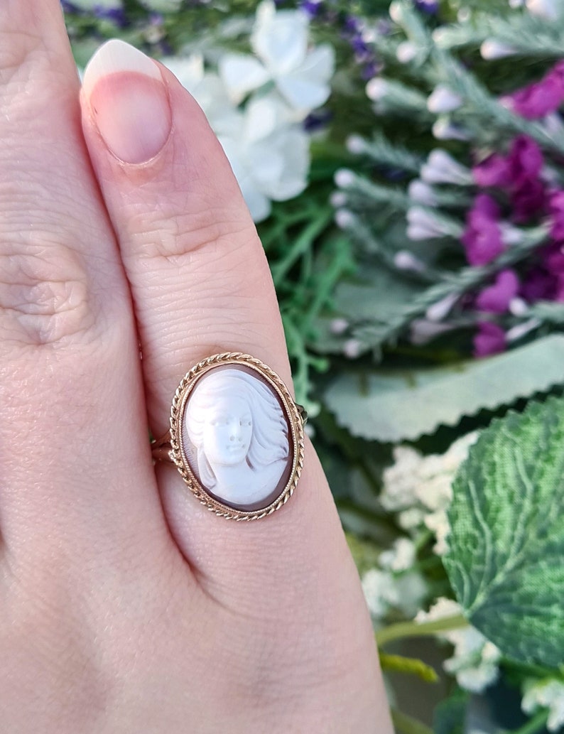 Op de afbeelding: Een gouden ring met een cameo met het profiel van een vrouw in wit op een bruine achtergrond.
