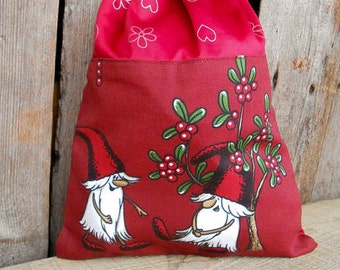 May include: Red drawstring bag with a white and red floral pattern on the top and a red background with two white gnomes and a green bush with red berries on the bottom.