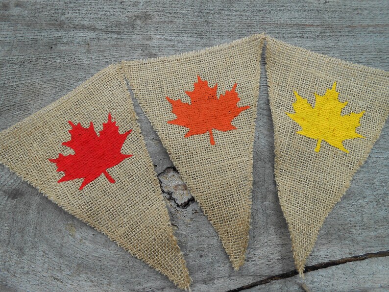 May include: Three burlap pennant flags with painted maple leaf designs. The leaves are red, orange, and yellow.