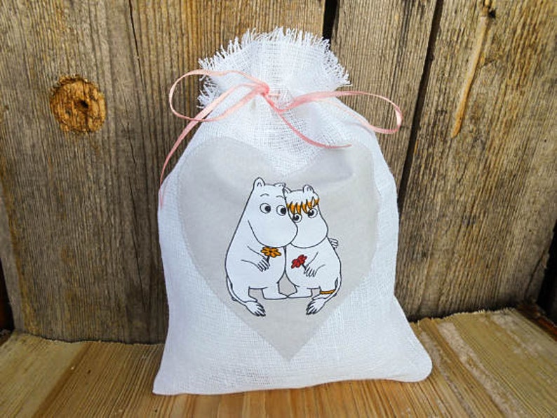 May include: A white linen drawstring bag with a pink ribbon and a heart-shaped window featuring a black and white illustration of two cartoon characters holding a flower.