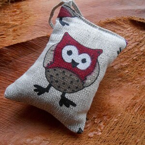May include: A small, gray linen owl-shaped pillow with a red and white owl design. The owl has black eyes and a brown and white patterned body.