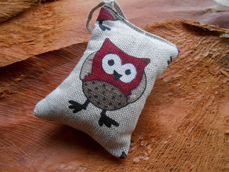 May include: A small, gray linen owl-shaped pillow with a red and white owl design. The owl has black eyes and a brown and white patterned body.