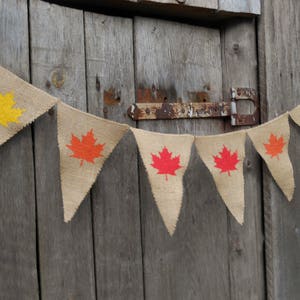 May include: A burlap banner with a string of pennants featuring orange, red, and yellow maple leaf designs. The banner is hanging on a wooden door.