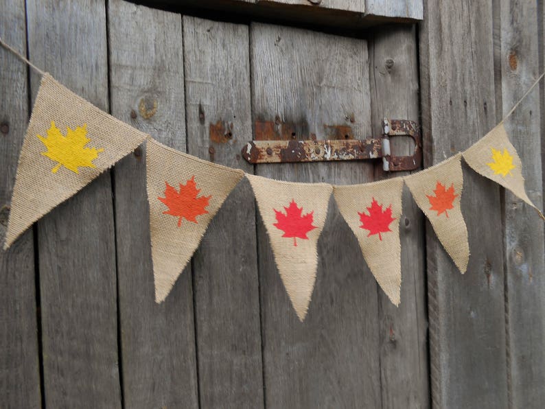 May include: A burlap banner with a string of pennants featuring orange, red, and yellow maple leaf designs. The banner is hanging on a wooden door.