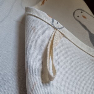 Puede incluir: Primer plano de una tela blanca doblada con un lazo. La tela presenta un patr&oacute;n repetido de p&aacute;jaros grises y naranjas. El lazo est&aacute; hecho de un material tejido de color crema, sujeto al borde de la tela.