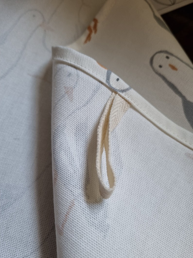 Puede incluir: Primer plano de una tela blanca doblada con un lazo. La tela presenta un patr&oacute;n repetido de p&aacute;jaros grises y naranjas. El lazo est&aacute; hecho de un material tejido de color crema, sujeto al borde de la tela.