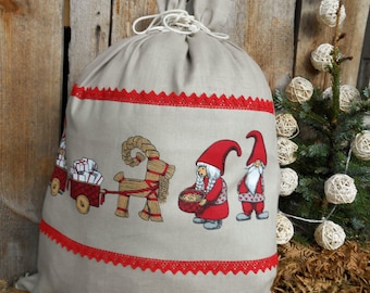 May include: A beige linen sack with a red and white trim. The sack features a festive scene with a gnome pulling a cart full of presents, a gnome holding a bowl of food, and a straw goat.