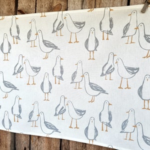 Puede incluir: Tela blanca con un patr&oacute;n repetido de gaviotas grises y beige. Las gaviotas est&aacute;n dibujadas en un estilo simple e ilustrativo. La tela cuelga sobre un fondo de madera.