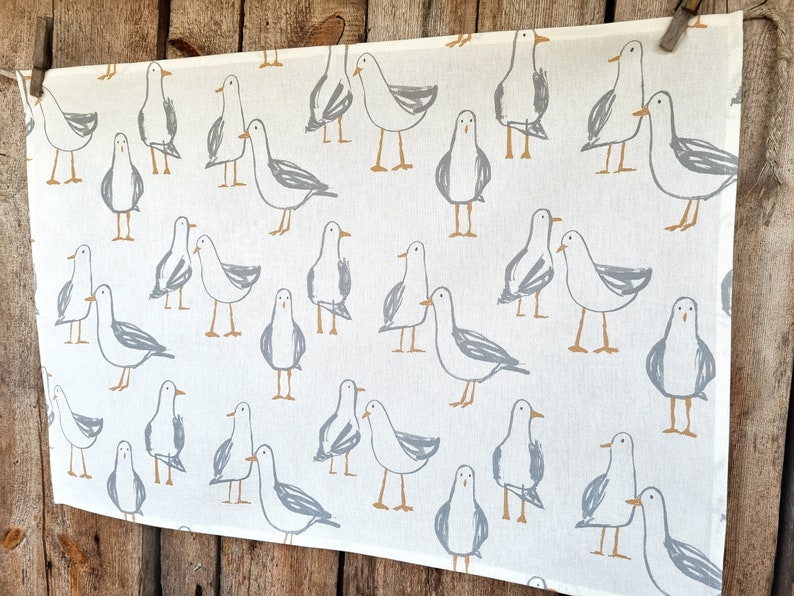 Puede incluir: Tela blanca con un patr&oacute;n repetido de gaviotas grises y beige. Las gaviotas est&aacute;n dibujadas en un estilo simple e ilustrativo. La tela cuelga sobre un fondo de madera.