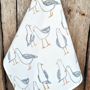 Puede incluir: Pa&ntilde;o de cocina blanco con un patr&oacute;n repetido de gaviotas grises estilizadas con patas y picos marrones. El pa&ntilde;o est&aacute; doblado y colgado sobre un fondo de madera r&uacute;stica. El dise&ntilde;o es sencillo y moderno.