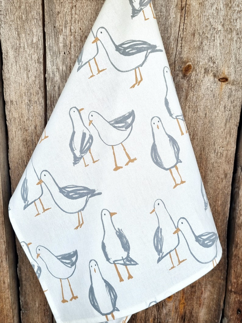 Puede incluir: Pa&ntilde;o de cocina blanco con un patr&oacute;n repetido de gaviotas grises estilizadas con patas y picos marrones. El pa&ntilde;o est&aacute; doblado y colgado sobre un fondo de madera r&uacute;stica. El dise&ntilde;o es sencillo y moderno.