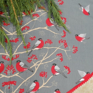 May include: A gray and white fabric Christmas tree skirt with a red trim. The skirt features a repeating pattern of red berries, branches, and red birds with white accents.