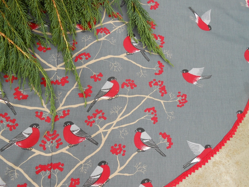 May include: A gray and white fabric Christmas tree skirt with a red trim. The skirt features a repeating pattern of red berries, branches, and red birds with white accents.