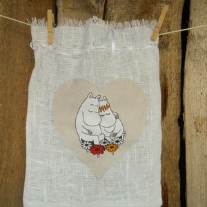 May include: White linen drawstring bag with a heart-shaped patch featuring two cartoon characters hugging. The characters are white with black outlines and are surrounded by colorful flowers.
