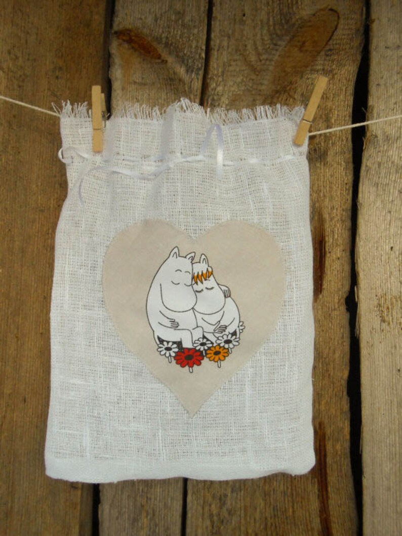 May include: White linen drawstring bag with a heart-shaped patch featuring two cartoon characters hugging. The characters are white with black outlines and are surrounded by colorful flowers.