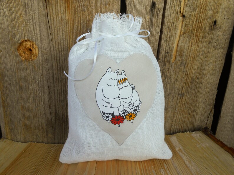 May include: White linen drawstring bag with a heart-shaped patch featuring a cartoon illustration of two white Moomins hugging. The Moomins are surrounded by red, orange, and yellow flowers.
