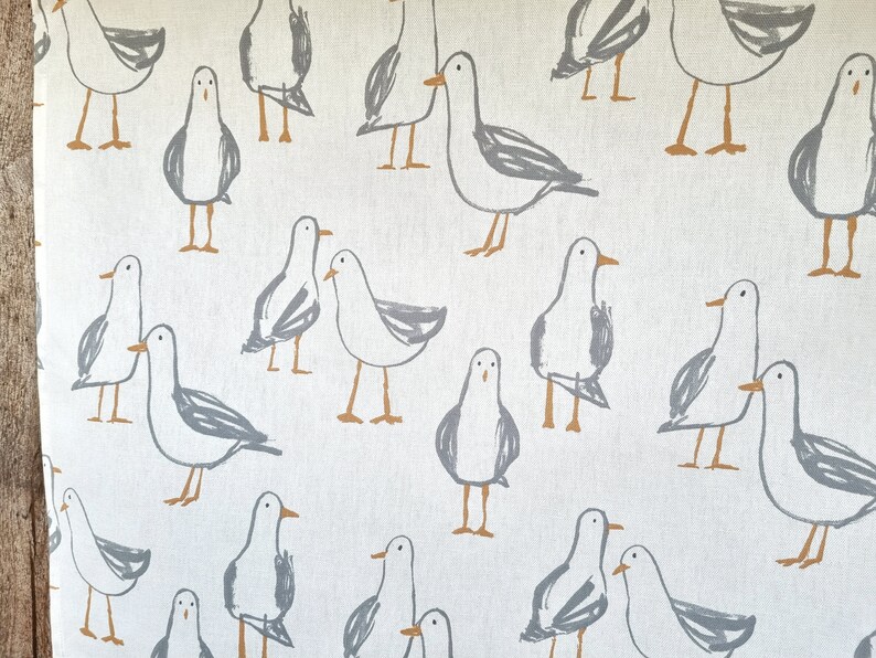 Puede incluir: Tela blanquecina con un patr&oacute;n repetido de gaviotas grises estilizadas con patas y picos naranjas. Las gaviotas est&aacute;n en varias poses, creando un dise&ntilde;o caprichoso y n&aacute;utico. La tela parece ser de algod&oacute;n o lino.