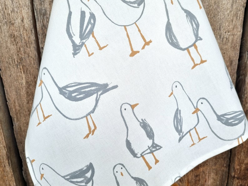 Puede incluir: Tela blanca con un patr&oacute;n repetido de gaviotas estilizadas en gris y marr&oacute;n. Las gaviotas est&aacute;n representadas con dibujos de l&iacute;neas simples, y la tela tiene un aspecto ligeramente texturizado. El dise&ntilde;o evoca un tema costero.