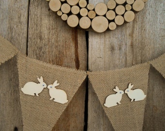 May include: A burlap banner with white wooden bunny cutouts. The banner is hanging on a rustic wooden background with a wood slice wreath above it.