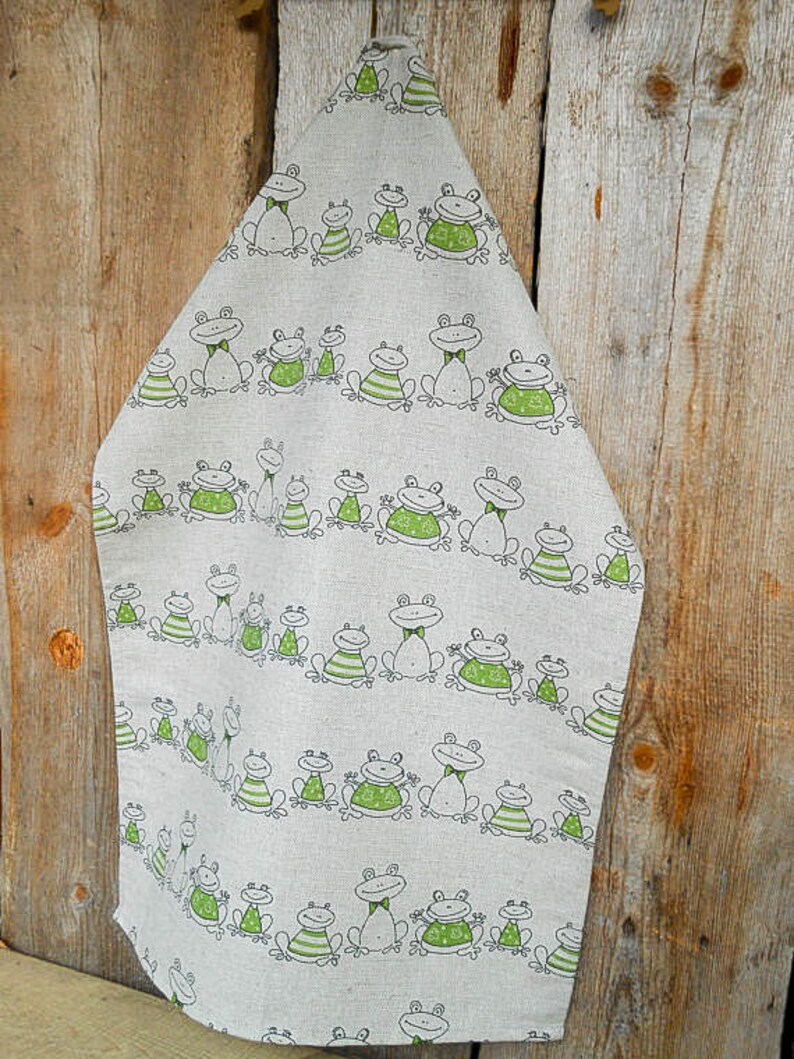 Puede incluir: Un pa&ntilde;o de cocina de lino con un patr&oacute;n de ranas verde y blanco. Las ranas est&aacute;n en diferentes poses y tienen manchas verdes. El pa&ntilde;o est&aacute; colgado en una superficie de madera.