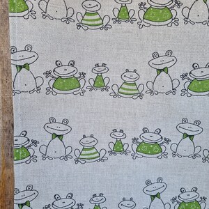 Puede incluir: Un pa&ntilde;o de cocina de tela blanca con un patr&oacute;n repetido de ranas verdes con contornos negros. Las ranas llevan pajaritas y algunas tienen rayas en el vientre.