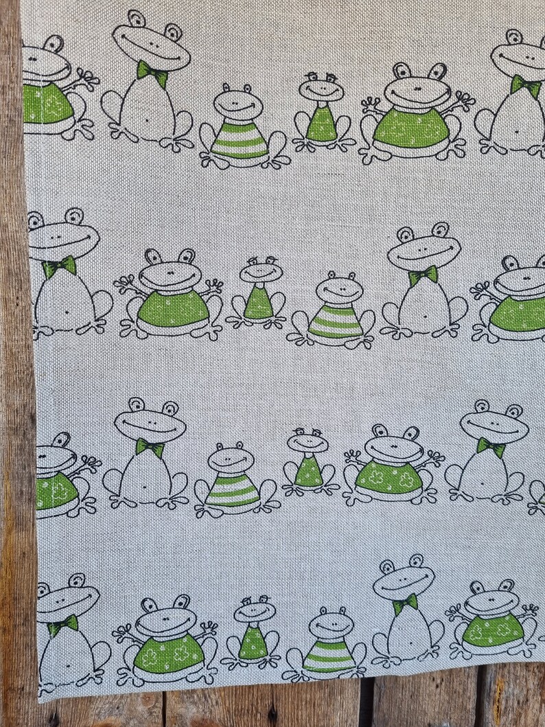 Puede incluir: Un pa&ntilde;o de cocina de tela blanca con un patr&oacute;n repetido de ranas verdes con contornos negros. Las ranas llevan pajaritas y algunas tienen rayas en el vientre.