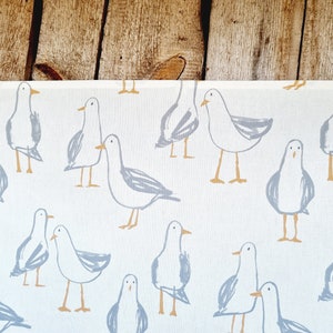 Puede incluir: Tela blanca con un patr&oacute;n repetido de gaviotas de dibujos animados en gris y beige. Las gaviotas est&aacute;n dibujadas con l&iacute;neas y formas simples, lo que les da una apariencia caprichosa. El fondo es de color crema claro.