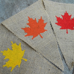 May include: Three burlap pennants with painted maple leaf designs. The leaves are orange, red, and yellow.