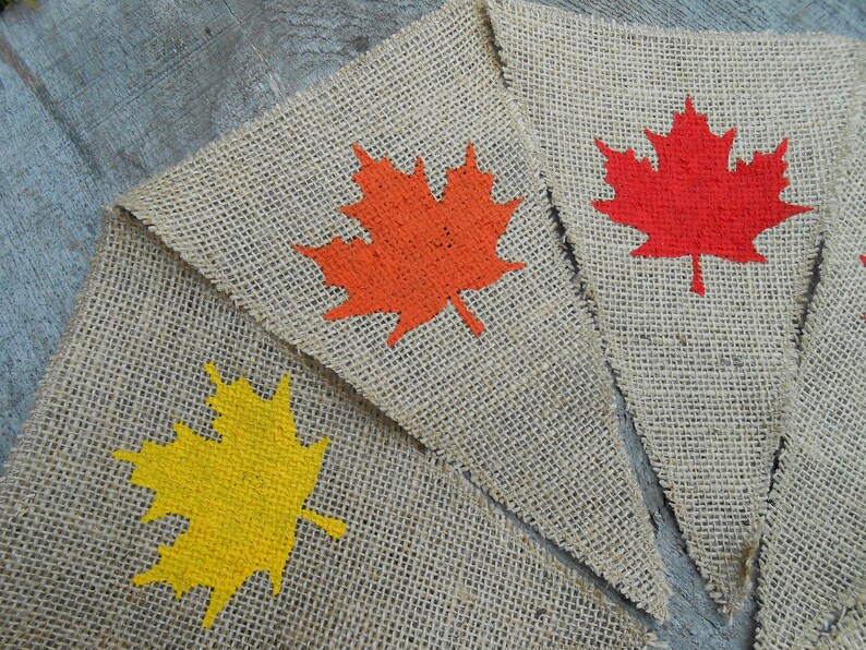 May include: Three burlap pennants with painted maple leaf designs. The leaves are orange, red, and yellow.
